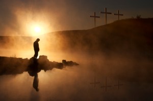 Dramatic religious photo illustration of Good Friday and Easter Sunday Morning reflecting a prayerful moment of silence with a silhoutted person bowing his head, a warm sunrise rises over a foggy lake, and three crosses on a hill reflected in the water as well.